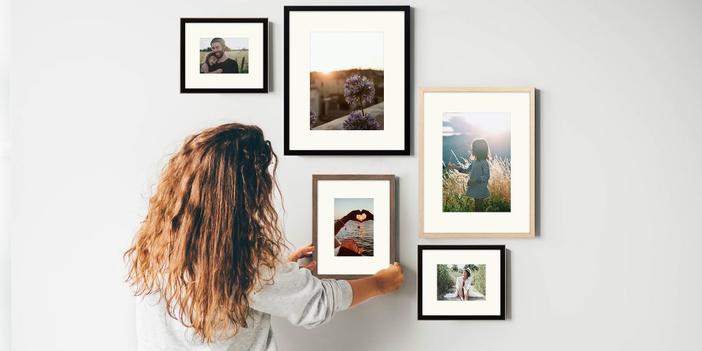 Woman with back facing camera hanging framed photographs on light gray wall.